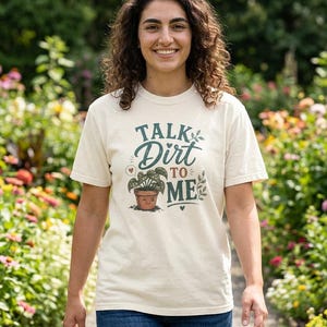 May include: Cream-colored t-shirt with the text "Talk Dirt to Me" in teal script and a graphic of a potted plant. The shirt is worn by a person in a garden setting, with colorful flowers in the background.