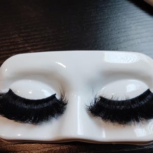 May include: A pair of dense, black false eyelashes displayed on a smooth, white, molded plastic stand shaped like closed eyelids. The lashes feature a full, voluminous design with fine, layered fibers that extend outward from a dark, flexible base.
