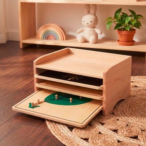 May include: Wooden Montessori-style learning toy with a natural finish. The toy features a multi-layered design with a tray and several wooden puzzles, including a tree and a sun. A wooden shelf with a rainbow and a stuffed bunny are in the background.
