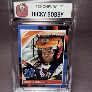 May include: A graded trading card featuring Ricky Bobby, driver of the #26 Chevrolet. The card is encased in a clear plastic holder and includes the text "RATED ROOKIE". The card shows Bobby in his racing helmet.