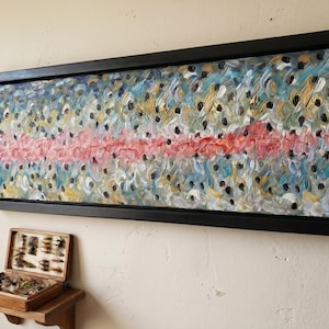 May include: Abstract oil painting of a fish in a black frame. The artwork features a central pink stripe with blue, yellow, and white brushstrokes, and black spots. A wooden fly fishing box sits below.