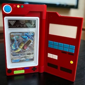 May include: Red card holder with a graded Pok&eacute;mon card featuring Gyarados. The card is encased in a clear plastic holder and is graded a 9. The card holder has a design with buttons and a screen, resembling a game console.
