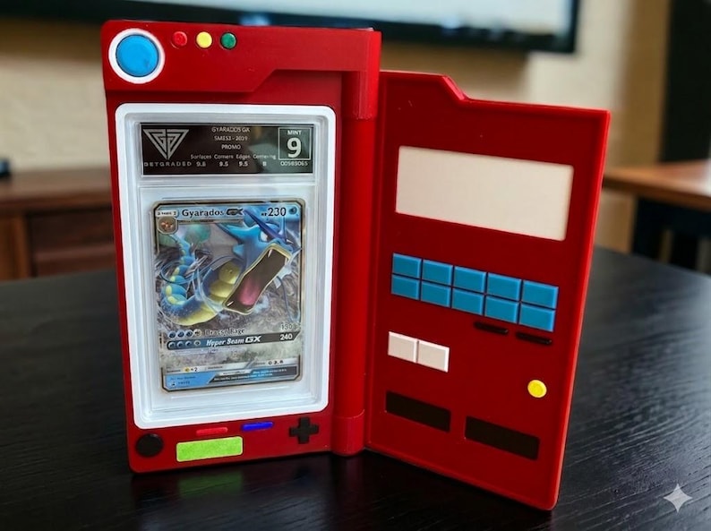 May include: Red card holder with a graded Pok&eacute;mon card featuring Gyarados. The card is encased in a clear plastic holder and is graded a 9. The card holder has a design with buttons and a screen, resembling a game console.