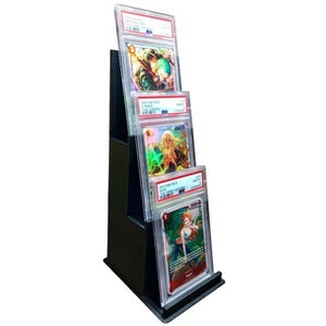 May include: A black, tiered display stand holding three graded trading cards. The cards feature colorful illustrations and text, encased in clear plastic holders. The stand is angled, allowing for easy viewing of the cards.
