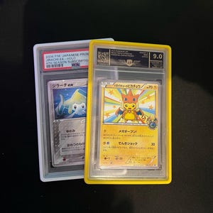 May include: Two graded Pok&eacute;mon trading cards. One card features a Jirachi EX-HOLO, and the other showcases a Pikachu character. The Pikachu card is graded 9.0. Both cards are encased in protective holders, with Japanese text visible.