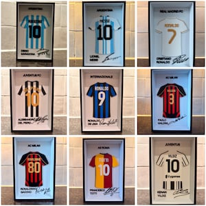 Può includere: Nove maglie da calcio incorniciate, ognuna con il nome, il numero e la squadra di un giocatore. Le maglie sono di vari colori, tra cui blu, bianco, rosso e nero. Le firme sono visibili sotto. Le cornici sono nere.
