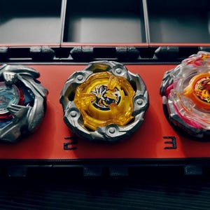 May include: Three colorful spinning tops in a red and black storage case. The tops have silver, yellow, and clear plastic components with intricate designs. The case is divided into compartments, with the number "2" and "3" visible.
