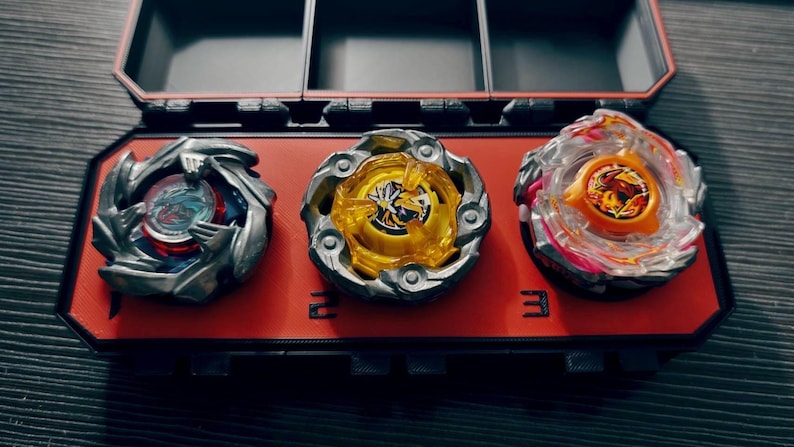 May include: Three colorful spinning tops in a red and black storage case. The tops have silver, yellow, and clear plastic components with intricate designs. The case is divided into compartments, with the number "2" and "3" visible.