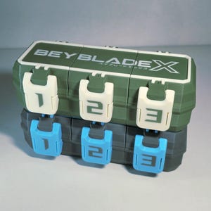 May include: A green and white plastic storage container with the text "BEYBLADE X" on top. The container has three white latches with the numbers 1, 2, and 3. Below are three blue latches with the same numbers.
