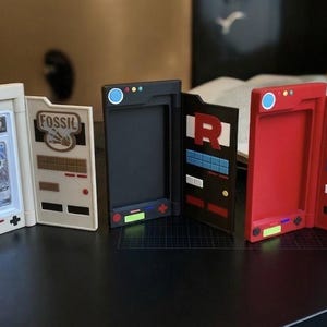 May include: A collection of open card holders in various colors, including white, black, and red. The holders feature designs and text, with one displaying a graded card. The holders are designed to resemble retro gaming devices.
