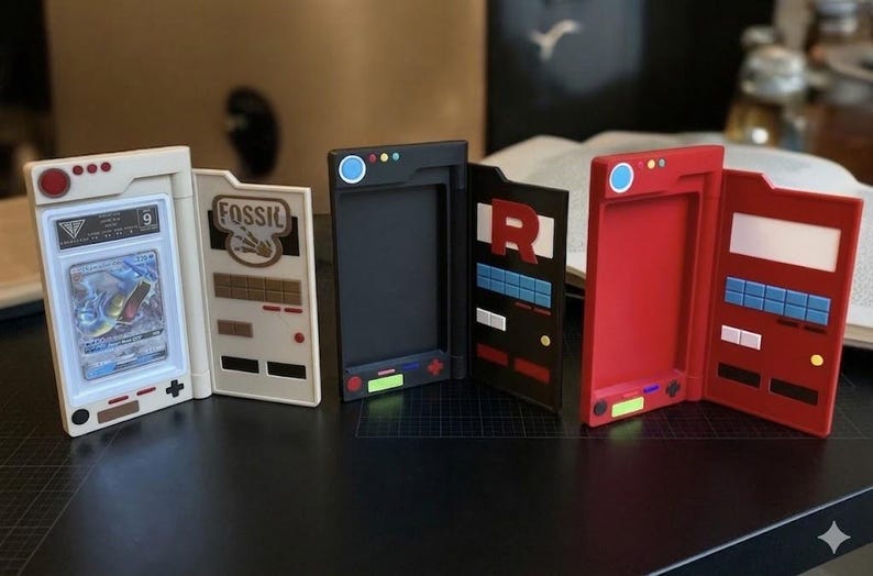May include: A collection of open card holders in various colors, including white, black, and red. The holders feature designs and text, with one displaying a graded card. The holders are designed to resemble retro gaming devices.