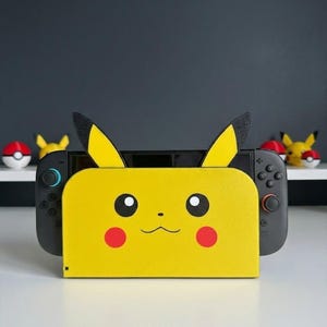 Könnte beinhalten: Ein gelbes Nintendo Switch-Dock im Pikachu-Design. Das Dock zeigt Pikachus Gesicht mit schwarzen Augen, roten Wangen und einem kleinen Mund. Die Ohren sind schwarz gespitzt. Die Switch-Konsole ist im Dock sichtbar, mit schwarzen Controllern auf jeder Seite.
