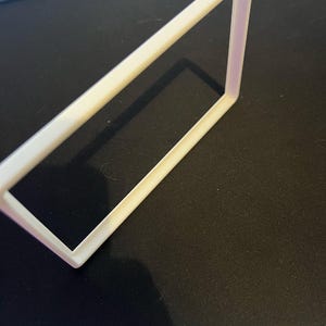 May include: A white rectangular frame with a slightly rounded edge. The frame is made of a solid material and casts a shadow on the black surface it rests upon. The frame is angled, showing its three-dimensional form.