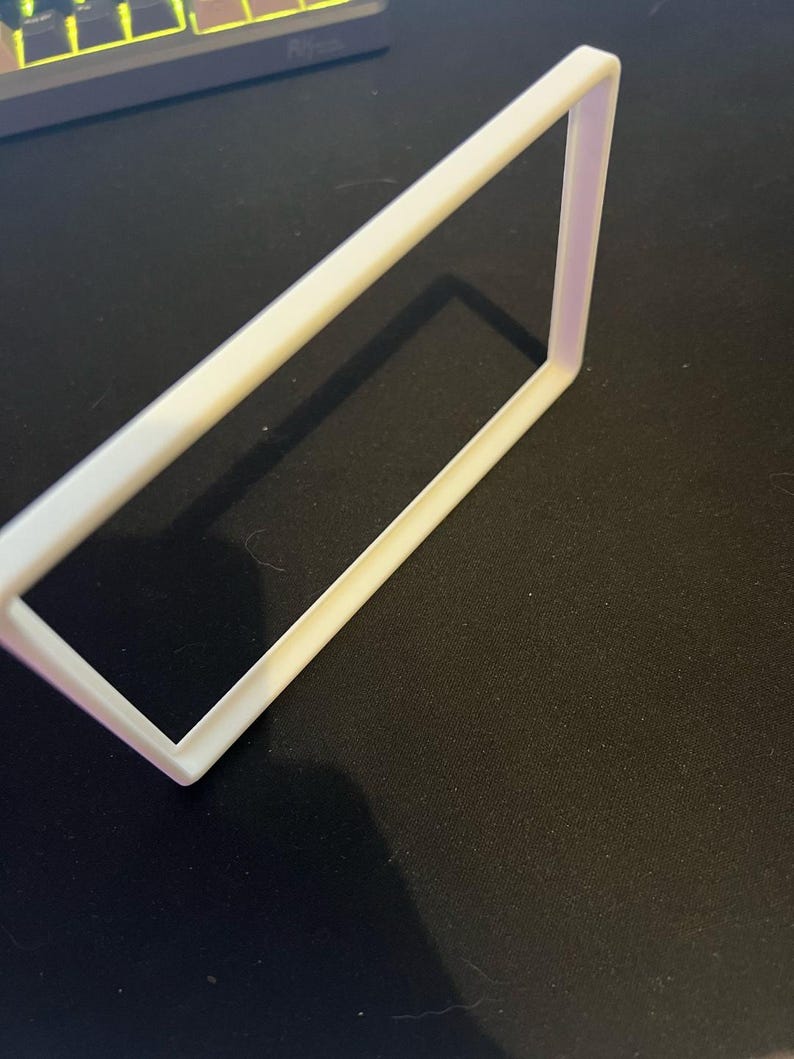 May include: A white rectangular frame with a slightly rounded edge. The frame is made of a solid material and casts a shadow on the black surface it rests upon. The frame is angled, showing its three-dimensional form.