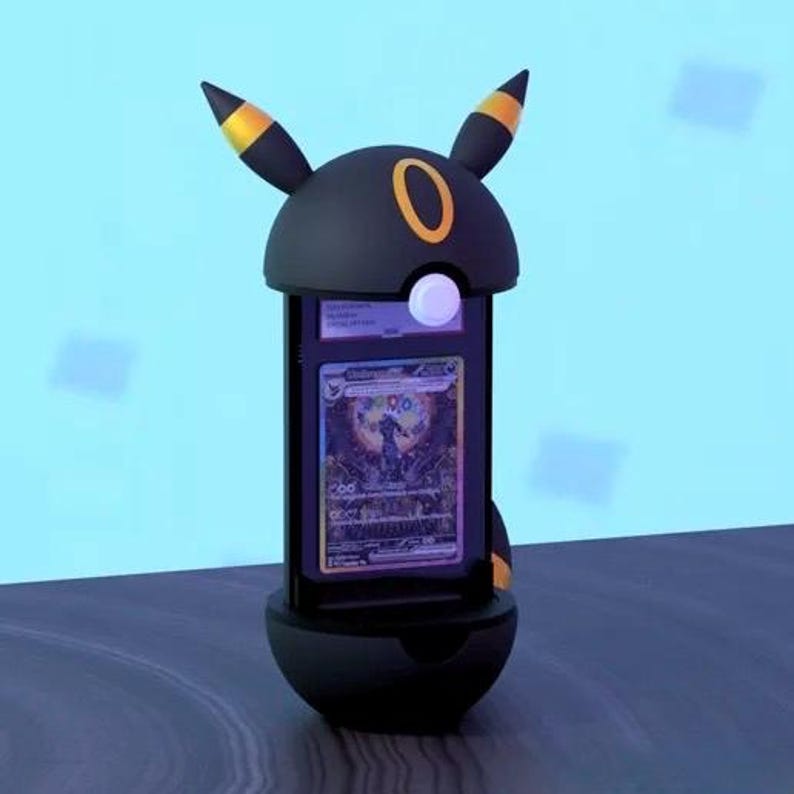 K&ouml;nnte beinhalten: Ein schwarzes Display im Umbreon-Pok&eacute;mon-Charakter-Design mit gelben Akzenten an den Ohren und einem goldenen Ring. Das Display enth&auml;lt eine Pok&eacute;mon-Sammelkarte mit einer farbenfrohen Illustration. Der Hintergrund ist hellblau.