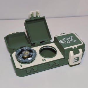 May include: A green and white toy storage case with a spinning top inside. The case has a hinged lid and a compartment for the toy. The case has the number 3 and the letters EX printed on it.
