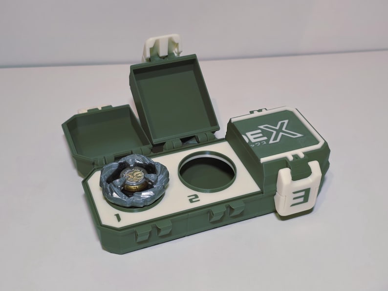 May include: A green and white toy storage case with a spinning top inside. The case has a hinged lid and a compartment for the toy. The case has the number 3 and the letters EX printed on it.