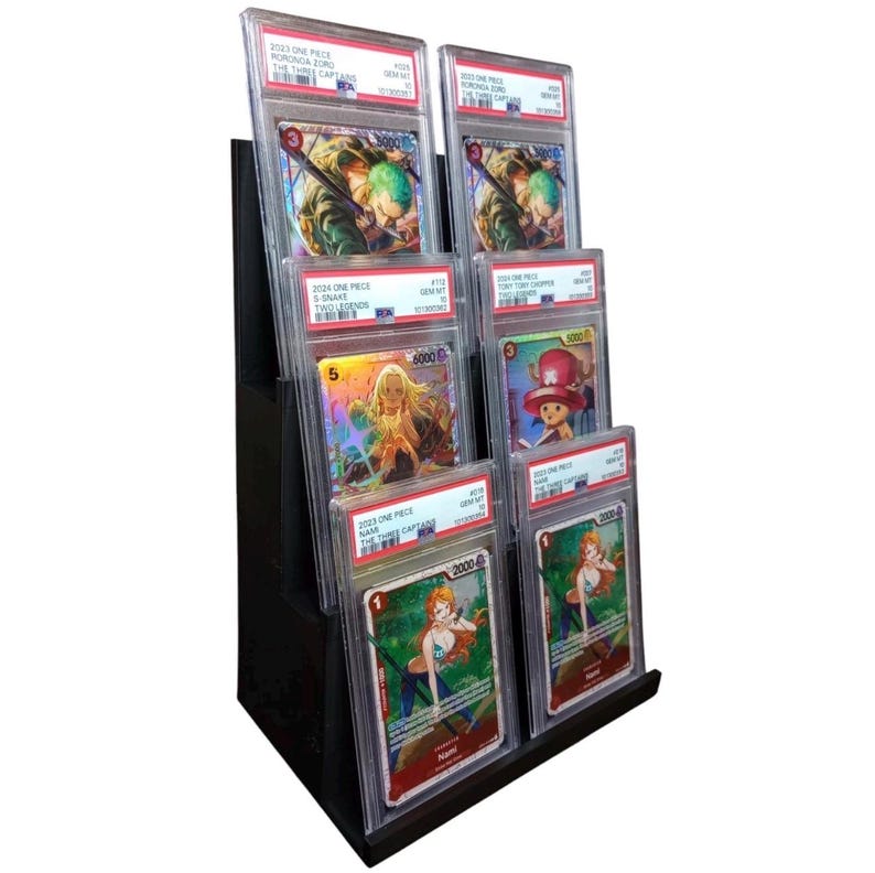 May include: A black display stand holding multiple graded trading cards. The cards feature colorful illustrations of anime characters, including Roronoa Zoro and Nami, from the One Piece series. The cards are encased in protective holders.