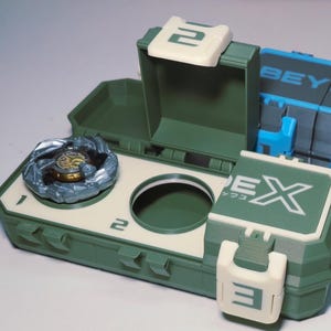 May include: A green and off-white toy storage case with a spinning top inside. The case has a hinged lid and a circular opening. The word "EX" and the number "2" are visible on the case.