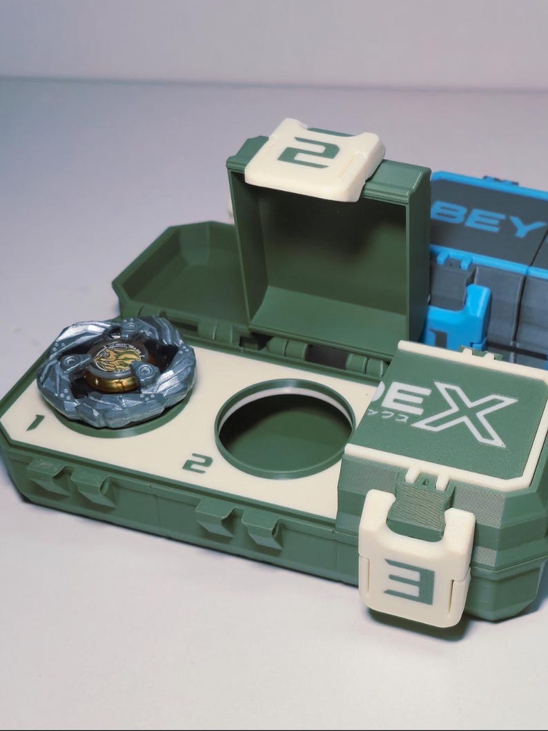 May include: A green and off-white toy storage case with a spinning top inside. The case has a hinged lid and a circular opening. The word "EX" and the number "2" are visible on the case.