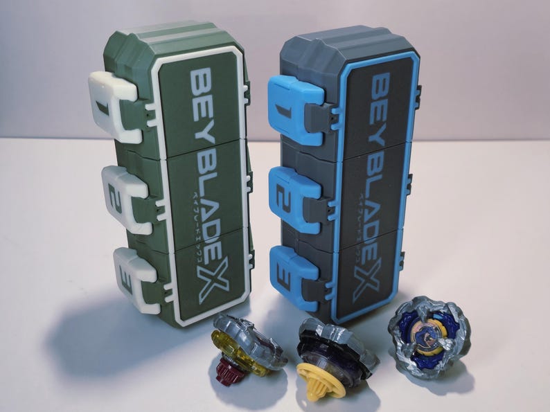 May include: Two toy storage cases and three spinning tops. The cases are green and gray with the text "BEYBLADE X" and Japanese text. The spinning tops are various colors, including yellow, purple, and blue.