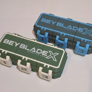 May include: Two rectangular storage cases with the text "BEYBLADE X" in white and Japanese characters. One case is green with white accents, the other is blue-gray with blue accents. Each case has three numbered latches.
