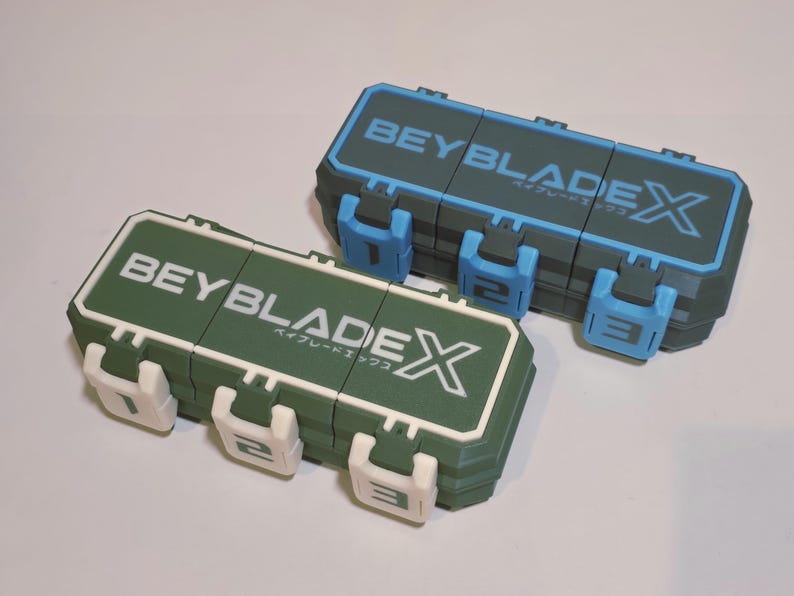 May include: Two rectangular storage cases with the text "BEYBLADE X" in white and Japanese characters. One case is green with white accents, the other is blue-gray with blue accents. Each case has three numbered latches.