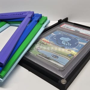 Op de afbeelding: Een stapel kleurrijke fotolijsten in blauwe, groene en paarse tinten, naast een zwart frame met een ruilkaart. De kaart toont een stripfiguur in een blauw en wit ontwerp.