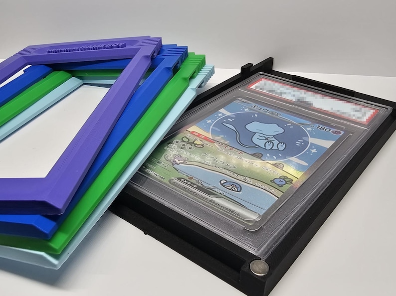 Op de afbeelding: Een stapel kleurrijke fotolijsten in blauwe, groene en paarse tinten, naast een zwart frame met een ruilkaart. De kaart toont een stripfiguur in een blauw en wit ontwerp.