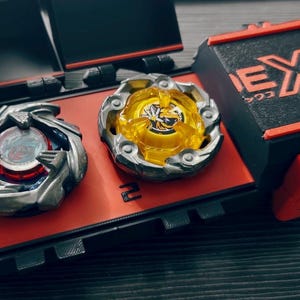 May include: A red and black carrying case with two spinning tops. One top is silver and red, the other is yellow and silver. The case has the word "EX" printed on it.