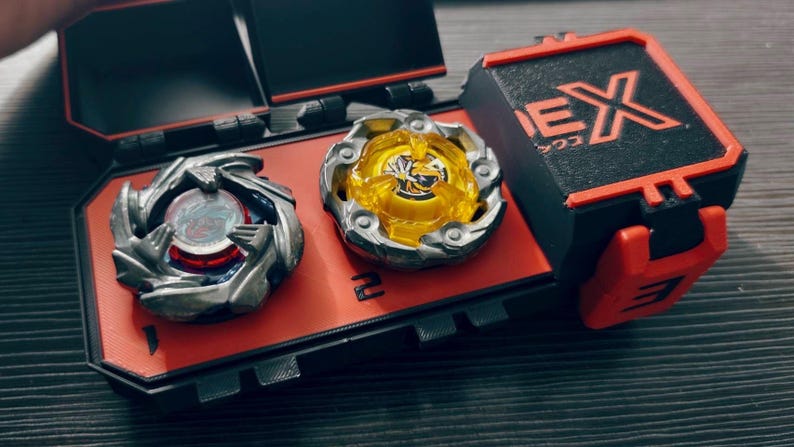 May include: A red and black carrying case with two spinning tops. One top is silver and red, the other is yellow and silver. The case has the word "EX" printed on it.