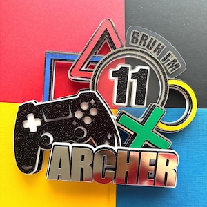 May include: A layered cake topper with a gaming theme. It features a black glitter controller, the name "ARCHER" in silver, and various game symbols. A circle with the number "11" is also included, along with the text "BRUH IM". The background is a mix of red, blue, yellow, and black.
