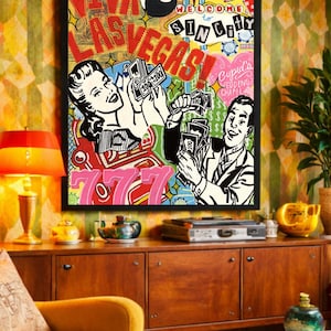 May include: A framed pop art style print with a black border, featuring the words "Viva Las Vegas!" and "Welcome to Sin City." The artwork depicts a woman and a man holding stacks of money, with casino-themed imagery and the number 777.