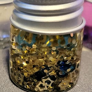 May include: A small glass jar with a silver lid filled with gold glitter, blue and black glitter, and star-shaped glitter. The jar is sitting on a gray surface.
