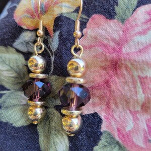 May include: A pair of dangle earrings featuring gold-tone metal hooks and spacers alternating with faceted dark purple crystal beads. Each earring consists of two round gold-tone beads and one central faceted purple bead, suspended from a gold-tone wire hook.