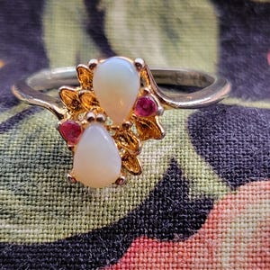 May include: A gold-toned ring featuring a cluster of pear-shaped, off-white gemstones and small red stones. The ring's design includes a floral-like setting, with the gemstones arranged on a band. The ring is set against a colorful floral fabric background.