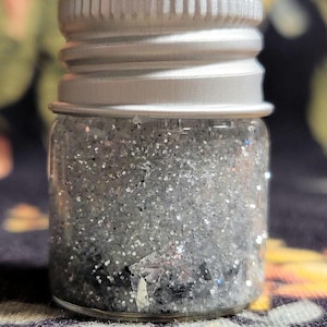 May include: A small, clear glass jar filled with silver glitter. The jar has a silver metal lid. The glitter appears to be a mix of fine and larger pieces, reflecting light. The jar is sitting on a patterned fabric.