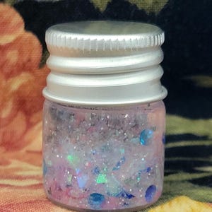May include: A small glass jar with a silver lid filled with pink, blue, and iridescent glitter. The glitter is suspended in a clear liquid. The jar is sitting on a floral patterned fabric.
