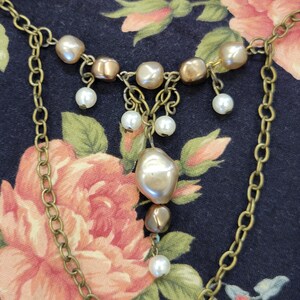 May include: A vintage-style necklace with a gold-tone chain and pearl accents. The necklace features a series of irregular-shaped pearls and smaller, round pearls that dangle from the chain. The background is a dark floral pattern.
