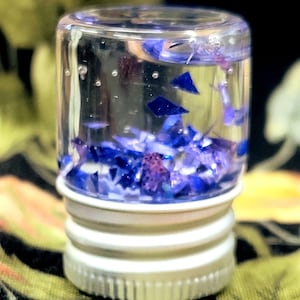 May include: A small, clear glass bottle with a silver cap, filled with clear liquid and blue and purple glitter. The bottle has a white base and is sitting on a patterned surface.