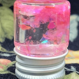 May include: A small, clear glass bottle filled with pink glitter and iridescent flakes. The bottle has a silver metal base and a clear, cylindrical body. The glitter and flakes are suspended in a clear liquid, creating a shimmering effect.