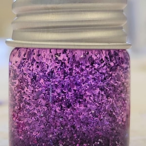 May include: A small glass jar filled with purple glitter. The glitter is a mix of shapes and sizes, creating a sparkling effect. The jar has a silver metal lid. The glitter appears to be for crafts or decorative purposes.