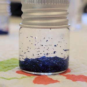 May include: A small, clear glass jar with a silver metal lid. The jar is filled with dark blue glitter. The glitter is concentrated at the bottom of the jar, with some particles suspended in the clear liquid.