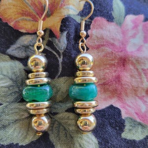 May include: A pair of dangle earrings featuring gold-toned metal hooks and a vertical arrangement of polished gold-toned spherical and disc-shaped beads, centered with a single irregular, smooth, vibrant green bead on each earring.