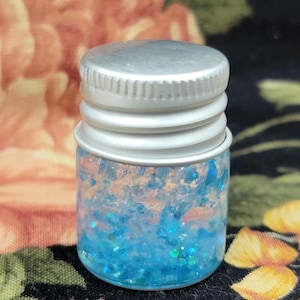 May include: A small, clear glass jar with a silver metal lid. The jar is filled with blue and iridescent glitter and small pieces of pink and orange material. The background is a floral pattern with pink and yellow flowers.