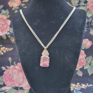 May include: A silver chain necklace with a small, clear glass bottle pendant filled with pink glitter. The bottle has a gold-colored cap and is displayed on a black mannequin.