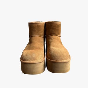 Könnte beinhalten: Ein Paar knöchelhohe Stiefel aus beigem Wildleder mit einer dicken, flachen Plateausohle. Die Stiefel haben eine abgerundete Zehenpartie, eine weich texturierte Oberfläche und ein sichtbares Plüschfutter am oberen Rand zum Hineinschlüpfen.
