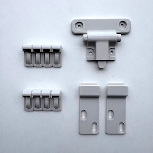 Può includere: Un set di componenti hardware in plastica grigio chiaro stampati in 3D, tra cui un meccanismo di chiusura a forma di T con fori per le viti, due piastre di montaggio rettangolari con asole ovali e otto piccoli fermagli a clip.