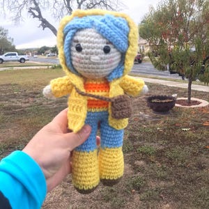 Peut inclure: Poup&eacute;e au crochet faite &agrave; la main avec une veste &agrave; capuche jaune, des cheveux bleus et un pantalon bleu avec des bottes jaunes. La poup&eacute;e a un petit sac marron et une chemise ray&eacute;e orange et jaune. La poup&eacute;e est tenue devant un arri&egrave;re-plan ext&eacute;rieur flou.