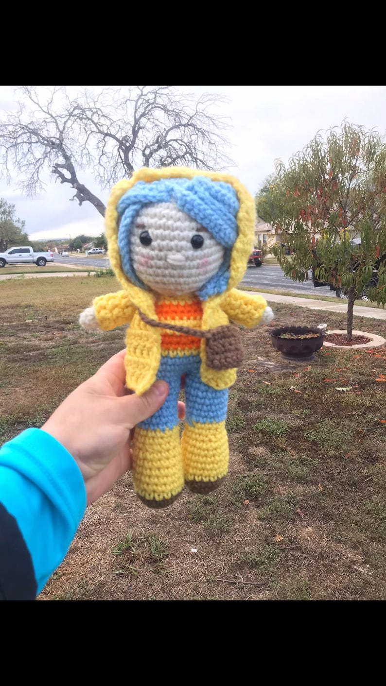 Peut inclure: Poup&eacute;e au crochet faite &agrave; la main avec une veste &agrave; capuche jaune, des cheveux bleus et un pantalon bleu avec des bottes jaunes. La poup&eacute;e a un petit sac marron et une chemise ray&eacute;e orange et jaune. La poup&eacute;e est tenue devant un arri&egrave;re-plan ext&eacute;rieur flou.